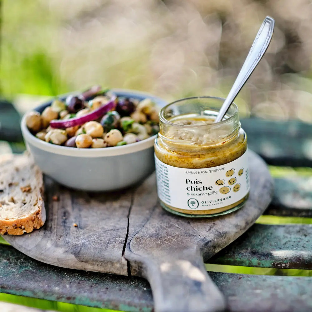 Sklenice 100g cizrnové pomazánky Oliviers&Co, s miskou cizrny a krajícem chleba na pozadí