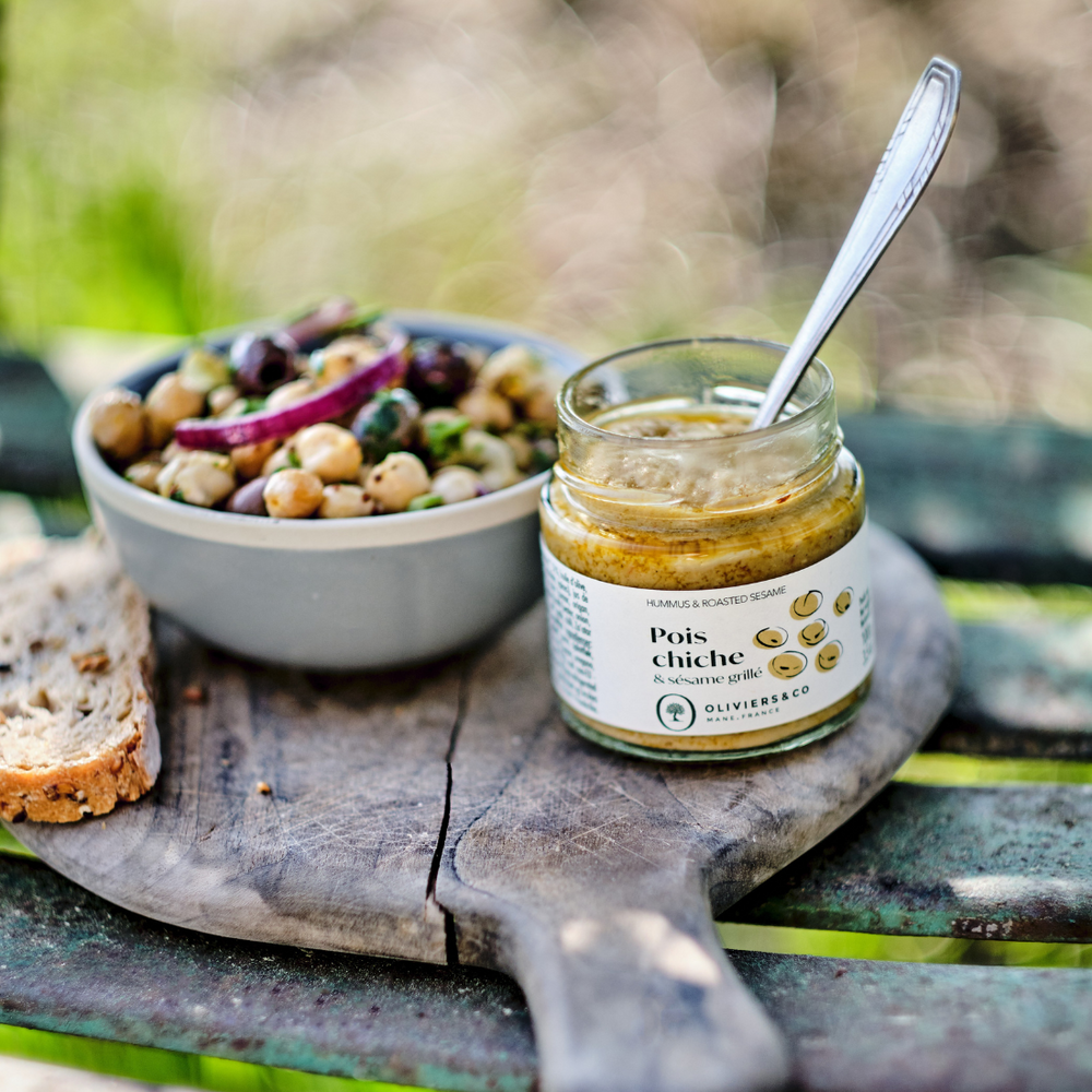 Sklenice 100g cizrnové pomazánky Oliviers&Co, s miskou cizrny a krajícem chleba na pozadí